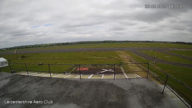 An outdoor view of an airfield or airport area with a paved runway in the distance. In the foreground, there is a balcony or observation deck with a black metal railing and painted markings on the ground. The sky is overcast with gray clouds, and the surrounding landscape is mostly flat and open. The timestamp in the top right corner reads "22-05-2025 14:00:09," and the bottom left corner has the text "Leicestershire Aero Club."