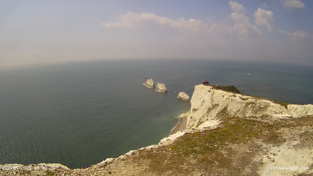 A high-angle view of a coastal landscape showing white chalk cliffs with green patches on top, extending into the sea. Several white rock formations rise from the water near the cliffs, and a small building or structure is visible at the edge of the cliffs. The sky is partly cloudy with patches of blue, and there is a sailboat in the distance on the water.