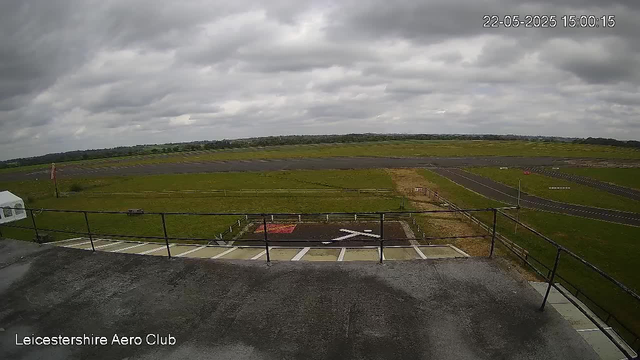 An outdoor scene at Leicester Airport on a cloudy day with dark gray clouds covering the sky. The runway and grassy areas stretch into the distance, and there is a helipad with a large white arrow painted on it in the foreground. A small portion of a building or platform with a black railing is visible at the bottom of the image. The timestamp in the top right corner indicates the date and time as May 22, 2025, at 15:00:15.