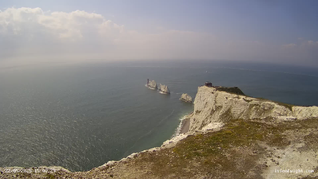 A panoramic view from a cliff overlooking the ocean with white chalk cliffs extending into the distance. There is a small structure on top of the cliff, and large rock formations rise from the sea nearby. The sky is partly cloudy with patches of sunlight. The scene appears calm and scenic.