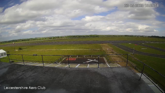 A wide view of an outdoor aircraft parking and runway area taken from a webcam, with a cloudy sky overhead and distant green fields. The foreground shows a balcony with a railing, and a marked helicopter landing pad is visible below. The image is labeled "Leicestershire Aero Club" in the bottom left corner, and a timestamp in the top right corner indicates the date and time as May 22, 2025, at 15:59:54.