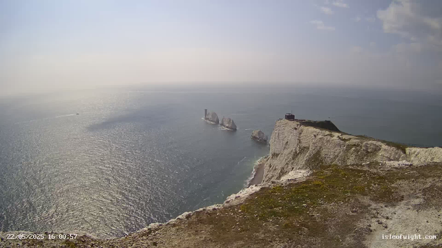 A view from a cliff overlooking the ocean, with white chalk cliffs and rock formations in the distance. The sky is partly cloudy and the water reflects the sunlight. There is a small structure or building on top of the cliff. The photo has a timestamp and website information at the bottom.