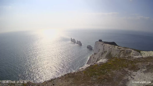 A view from a high cliff overlooking the ocean on a bright, sunny day. The sunlight reflects off the water, creating a shimmering effect. In the distance, there are several large sea stacks and a small lighthouse perched on the edge of the cliff. The foreground shows the rocky, grassy edge of the cliff.