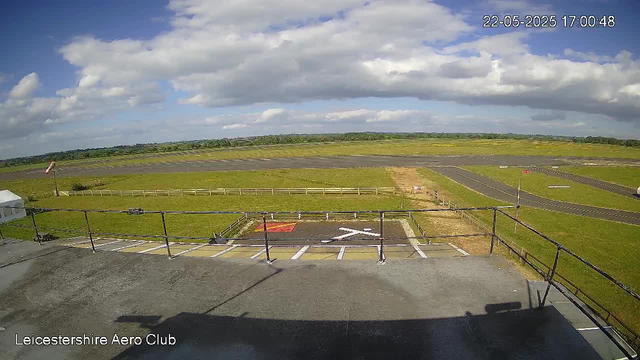 A view from a webcam overlooking an airfield with a runway in the distance, some taxiways, and grassy areas. The sky is partly cloudy with a mix of white clouds and blue sky. There is a small helipad or landing zone with markings in the foreground, and a parking space area below the webcam. The scene is captured during daytime, and the timestamp shows the date as May 22, 2025, at 5:00 PM. The location is identified as Leicesterhire Aero Club.
