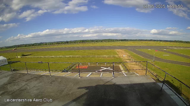 A view of an airfield from a webcam, showing a runway extension, grassy areas, and a small parking area in the foreground with marked parking spaces. The sky is partly cloudy with patches of blue, and the image is timestamped "22-05-2025 17:59:50." The location is identified as Leicestershire Aero Club.