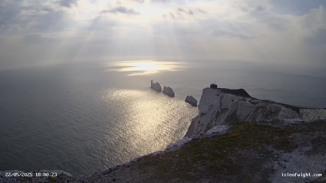 A view of white chalk cliffs overlooking the ocean during sunset or sunrise. Rocky rock formations protrude from the water near the cliffs, and a small building is visible on the edge of the cliff. The sky is partially cloudy, with rays of sunlight breaking through the clouds and reflecting off the water's surface. A timestamp at the bottom left indicates the date and time as 22/05/2025 at 18:00:23, and the website isleofwight.com is displayed at the bottom right.
