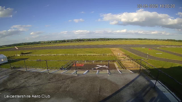 A view from a webcam at Leicesterhire Aero Club showing an open sky with scattered clouds. In the foreground, there is a parking area with white and blue parking lines, a railing, and a single vehicle partially visible on the left. Beyond the parking lot, a grassy field with a small paved section marked with a large white "X" is visible. Further in the distance, there is a paved runway with white markings and a red windsock on the right side. The horizon features a flat landscape with patches of trees and fields under a mostly clear sky with some fluffy clouds. The timestamp on the image reads "22-05-2025 19:00:20".