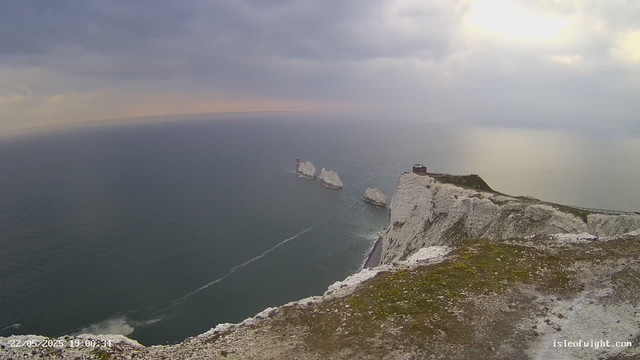 An aerial view of white chalk cliffs overlooking the ocean under a cloudy sky. A small building and a pathway run along the top of the cliffs, with several sea stacks and rocky formations extending into the water in the distance. The scene appears to be during late afternoon or early evening, with soft lighting and a calm sea.