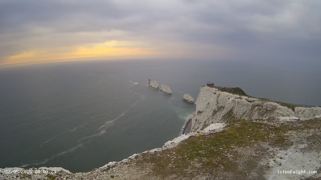 A view of the white chalk cliffs and rock formations along the coastline, with a small building and a railing on top, overlooking the ocean beneath a cloudy sky at sunset. The sea is calm with visible waves and a lighthouse can be seen on an island in the distance. The image has a timestamp of "22/05/2025 20:00:23" and the website "isleofwight.com" is visible in the bottom right corner.