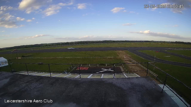 An outdoor view of a small airport or airfield: there is a runway with a parked airplane in the distance, surrounded by grassy fields, a fence, and a small helipad in the foreground. The sky is partly cloudy, and the image includes a timestamp in the top right corner showing "22-05-2025 20:00:45." The bottom left corner has the text "Leicestershire Aero Club."