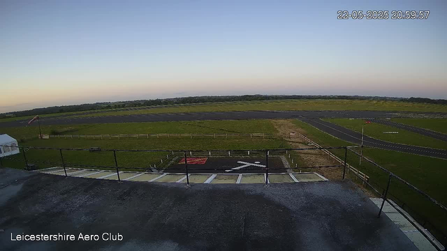 An empty helicopter landing pad with the letters "H" in white, surrounded by a black safety fence, at Leicestershire Aero Club. In the background, there are green fields and a small runway, with a clear blue sky and the late evening sunlight. The timestamp in the top right corner shows "22-05-2025 20:59:57."