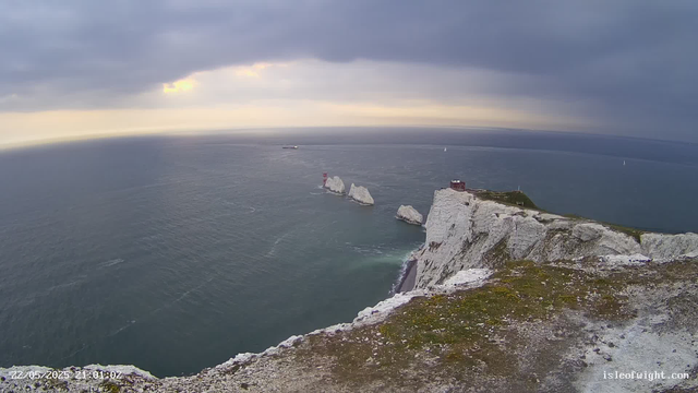 Aerial view of white chalk cliffs with patches of green grass, extending into the ocean under a cloudy sky. Several large white sea stacks rise from the water near the cliffs, and a small building is perched on top of the highest point of the cliffs. The ocean is calm with gentle waves, and a few sailboats and ships are visible in the distance. The timestamp at the bottom left indicates the date and time as 22/05/2025 at 1:01:02.