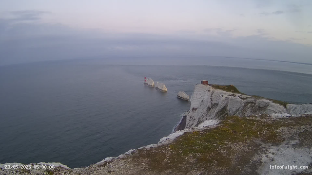 A view of white chalk cliffs overlooking the ocean during dawn or dusk. A small building and a lighthouse are visible on the cliff's edge, with a path running along the top. Large rocks are protruding from the water near the base of the cliffs, and the sky is partly cloudy with soft pastel colors.