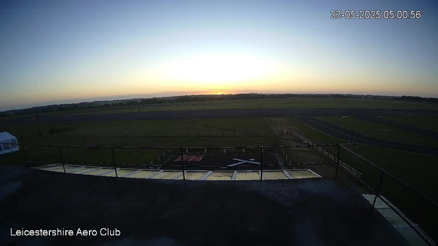 A wide-angle view from a webcam captures the early morning sky at sunrise over an open airfield. The horizon shows a faint glow of sunlight, with the sky transitioning from dark blue to lighter shades near the horizon. In the foreground, there is a rooftop with a metal railing around the edge, and a small helipad marked with a white "H" and an arrow. The bottom left corner has the text "Leicestershire Aero Club". The upper right corner displays the date and time as "23-05-2025 05:00:56".