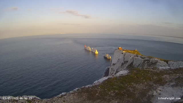 A view of white chalk cliffs with patches of green grass on top, overlooking the ocean. Several large rocks and sea stacks are visible offshore, with one prominent lighthouse standing on a small red structure. The sky is partly cloudy, and the scene is illuminated by warm sunlight, suggesting early morning or late afternoon. The timestamp shows "23/05/2025 06:00:49" and the website "isleofwight.com" is displayed in the bottom right corner.