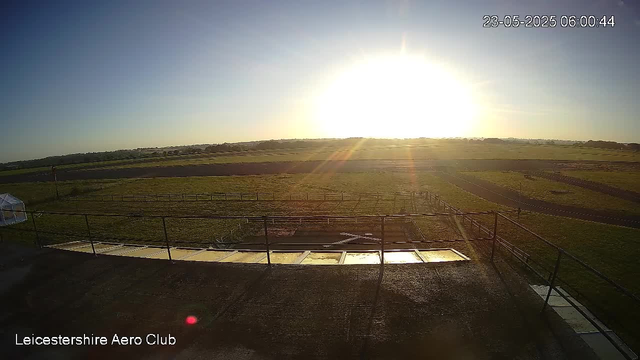 The image shows a wide outdoor view of a field at sunrise, with the bright sun near the horizon casting long shadows. There is a fenced area in the foreground and a small greenhouse on the left side. The landscape features open grassy fields with a clear sky overhead. The timestamp in the top right corner reads "23-05-2025 06:00:44," and the bottom left corner displays the text "Leicestershire Aero Club."