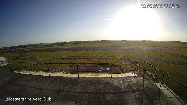 A wide view of an open grassy airfield with a runway, taken from an elevated perspective on a clear, sunny morning. The sky is mostly clear with the sun shining brightly in the upper right corner. In the foreground, there is a fenced area with some small structures or equipment. The bottom left corner has the text "Leicestershire Aero Club".