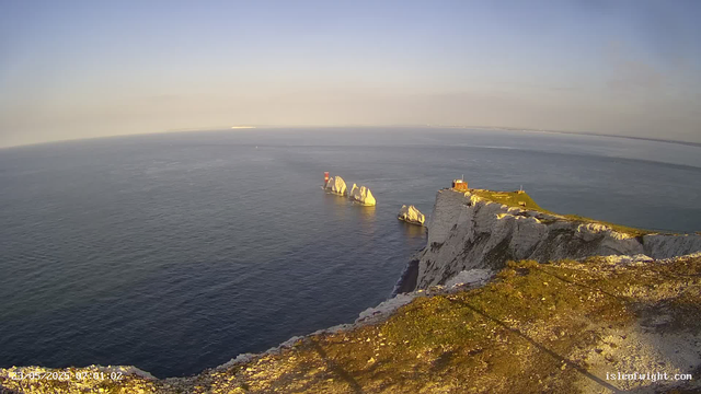 A view of white chalk cliffs with patches of green grass on top, jutting out into the calm, expansive sea. There is a lighthouse or small building on the edge of the cliff. The sky is clear with a soft gradient of light blue and the horizon stretches far into the distance. The photo appears to be taken during the early morning or late afternoon, suggested by the warm sunlight illuminating the scene. The timestamp indicates the image was captured on May 23, 2023, at 01:02.
