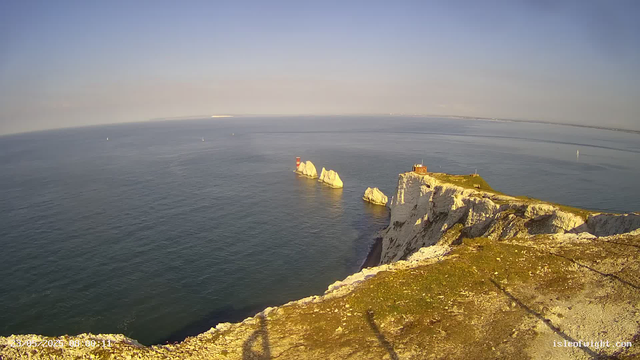 A view from a webcam showing white chalk cliffs with patches of green grass on top, overlooking the ocean. Several white chalk stacks rise from the water near the base of the cliffs. The sky is clear with a faint haze, and a few sailboats are visible in the distance on the calm sea. A lighthouse is situated on the edge of the cliff. The timestamp on the image reads "2023-05-20 08:00:11".
