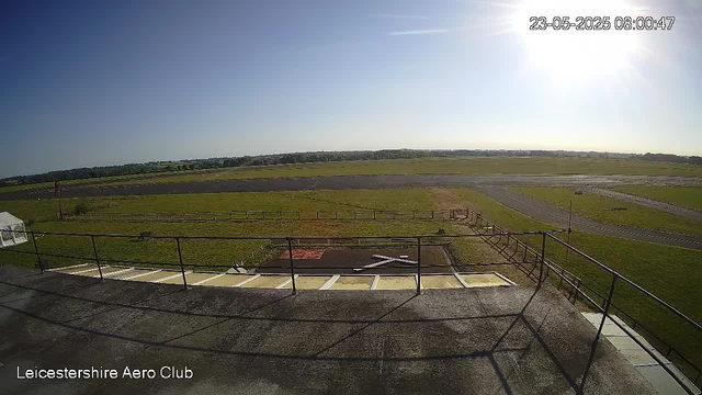 An outdoor scene of a runway area at Leicester Shire Aero Club, with a clear blue sky and the sun shining brightly in the upper right corner. The foreground shows a parking area with painted white lines and a metal railing, casting shadows on the ground. Beyond, there is a grassy field with pathways and a small fence. In the distance, the horizon features low hills and scattered buildings. The timestamp in the upper right corner reads "23-05-2023 08:00:47".