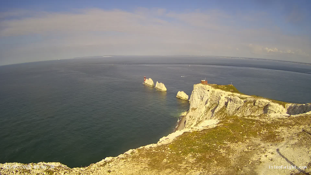 A view of white chalk cliffs overlooking the ocean, with several large rocks/sea stacks in the water nearby. On top of the cliffs, there is a small building and a lighthouse further out on a promontory. The sky is partly cloudy with some blue visible.