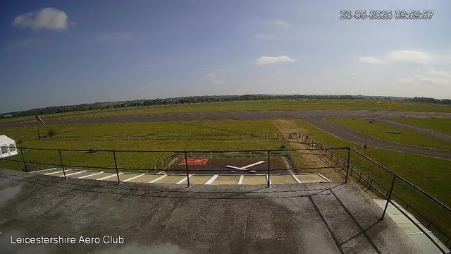 An outdoor view from a webcam at Leicestershire Aero Club shows a clear day with a blue sky and a few scattered clouds. In the foreground, there is a concrete area with a railing and a helipad marked with a white "H" on a black surface. Surrounding the helipad, there are grassy fields and a paved airstrip that extends into the distance. There are no aircraft or people visible in the scene. The timestamp in the upper right corner indicates the date and time as 23 May 2025, 09:59:57.