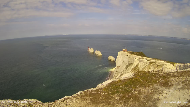 Aerial view of white chalk cliffs along the coastline with a small building on top, several large rocks near the shore, and a lighthouse. The ocean is calm with sailboats visible in the distance, under a partly cloudy sky.