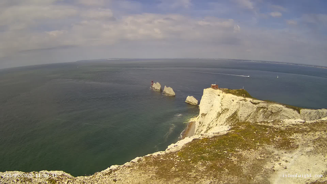 Aerial view of white chalk cliffs with a small building at the edge, overlooking the sea with several large rocks protruding from the water. The sky is partly cloudy, and sailboats are visible in the distance. A lighthouse stands on the cliff's edge.