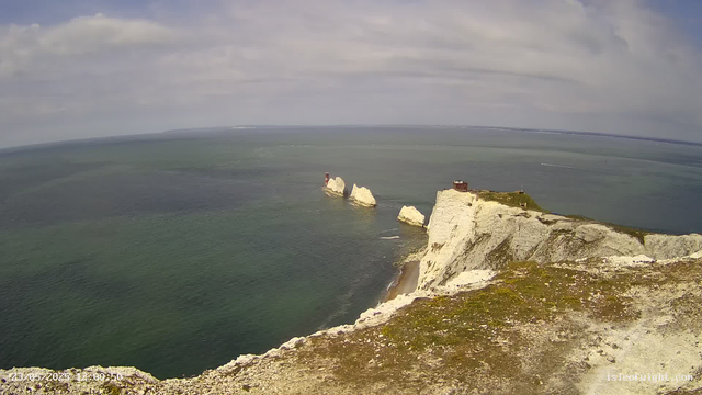 A panoramic view of white chalk cliffs rising sharply from the ocean, with a small building and a flagpole on top of the cliffs. Several large chalk stacks are visible offshore, with one featuring a lighthouse. The sea is a mix of green and blue hues, and the sky is partly cloudy with patches of sunlight. The image appears to be taken from a high vantage point or webcam, capturing a coastal landscape.