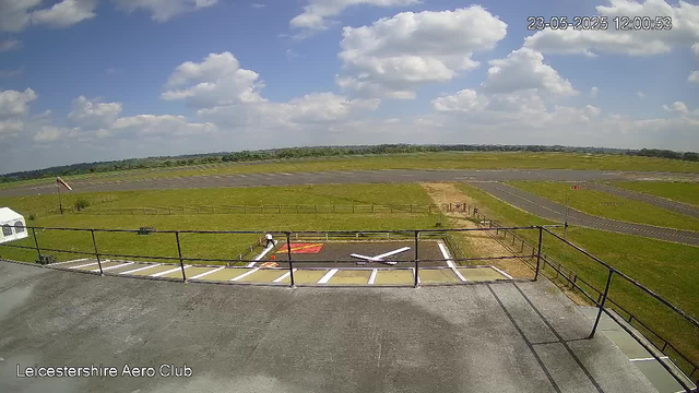A view from a webcam at Leicestershire Aero Club shows an airport runway and grassy areas under a partly cloudy sky. In the foreground, there is a rooftop with a metal railing, a white airplane model, and a red and yellow marking on the ground. The airport taxiway stretches into the distance, with some small buildings and trees visible on the horizon. The timestamp in the top right corner reads "23-05-2025 12:00:53".