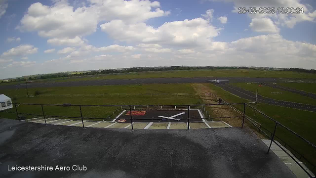 A view from a webcam at Leicestershire Aero Club shows a partly cloudy sky with scattered white clouds and blue patches. Below, there is a grassy airfield with a small helicopter on the helipad, surrounded by fencing and parking spaces. In the background, there are open fields and a distant horizon. The image is timestamped "23-05-2025 13:00:24."