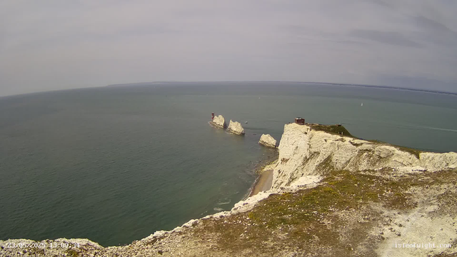 A panoramic view of white chalk cliffs along the coastline, with a grassy top and small structures on the edge. Several large rocky stacks are visible in the water offshore, and sailboats are scattered across the sea. The sky above is cloudy.
