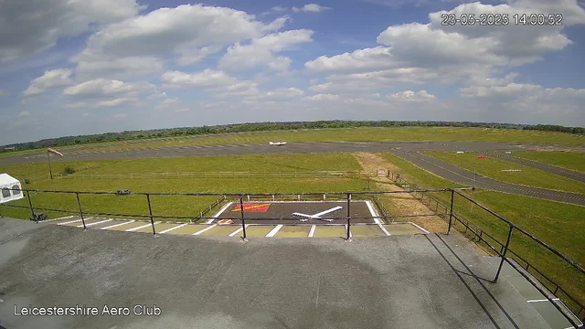 A view from a webcam overlooking Leicestershire Aero Club. In the foreground, there is a small parking area with marked parking spaces and a fenced helipad with a white helicopter-shaped symbol. A runway is visible in the middle ground, stretching across the landscape. In the background, there are open grassy fields and a sky filled with scattered clouds, with an aircraft taking off or landing on the runway. The timestamp in the upper right corner indicates the date and time as "23-05-2025 14:00:52".