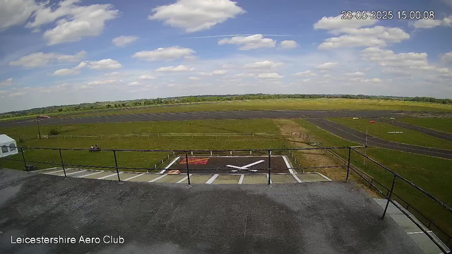 A view from a webcam at Leicestershire Aero Club showing a runway and airfield on a partly cloudy day. The sky is blue with scattered white clouds, and the horizon has distant green fields and trees. There is a small airplane symbol painted on a designated landing area, and the timestamp in the top right corner reads "23-05-2025 15:00:08."