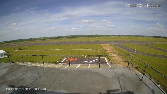 A view from a webcam at Leicesterwshire Aero Club showing a grassy airfield with runways in the distance, partly cloudy sky above, and a helicopter landing pad with white markings and a red area. The timestamp indicates the image was taken on May 23, 2025, at 16:00:32.