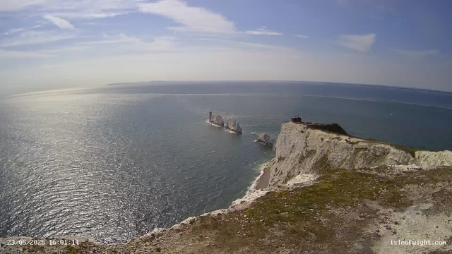 Aerial view of white chalk cliffs overlooking the ocean with several large rocks and sea stacks emerging from the water. The sky is partly cloudy with sunlight reflecting off the water's surface. A small building and a tall tower are situated on top of the cliffs. The horizon extends far into the distance, creating a scenic coastal landscape.