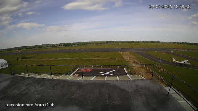 A view from a webcam showing an airport runway and taxiway, with a small airplane positioned on a helipad in the foreground. The sky is mostly clear with some scattered clouds. There is a grassy field surrounding the runway, and an American flag flying on a flagpole near the helipad. The image is timestamped "23-05-2025 16:59:52" and has a label "Leicestershire Aero Club" at the bottom left corner.