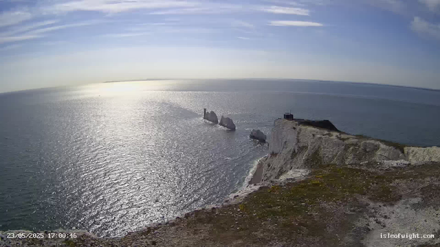A view from a webcam showing a bright, sunny day over a large body of water with white chalk cliffs in the foreground. The cliffs extend into the distance with some rock formations visible in the water nearby. The sky is mostly clear with a few wispy clouds. The camera timestamp indicates the date and time as May 23, 2025, at 5:00 PM.