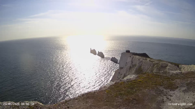 A view of a rocky coastline with white chalk cliffs overlooking the ocean. Several large rock formations are visible just off the coast, emerging from the water. The sun is low on the horizon, creating a bright glare that reflects on the surface of the sea. The sky is mostly clear with a few wispy clouds. The date and time stamp in the bottom left corner reads "23/05/2025 18:00:50," and the bottom right corner has the website "isleofwight.com."