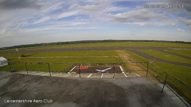 A view from a webcam at Leicesterhire Aero Club shows a clear sky with some scattered clouds. The scene includes a small parking area with a designated helicopter landing pad marked with a white "H" and red lines. The tarmac surface has white parking space lines and a fence surrounding the landing zone. In the background, there is a grassy field with a runway extending into the distance and some windsocks. The image is timestamped "23-05-2025 18:00:51" in the top right corner.