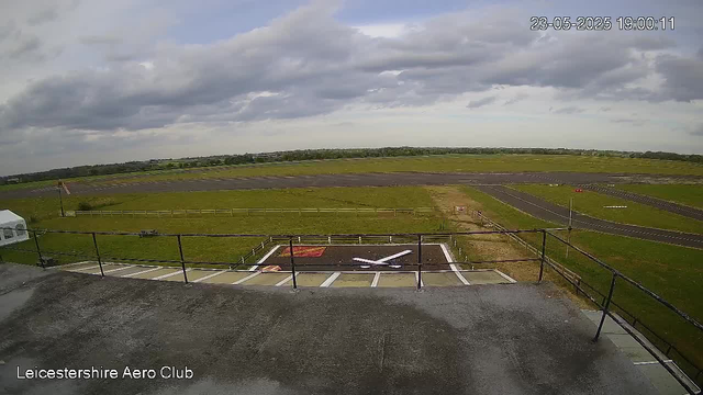 The image shows a view from a webcam overlooking an airfield. In the foreground, there is a small helicopter landing pad with a painted helicopter symbol and area markings, surrounded by a metal railing. Beyond the landing pad, there are grassy fields and a paved taxiway or runway stretching into the distance. The sky above is partly cloudy with patches of blue visible, and the time and date stamp in the upper right corner reads "23-05-2025 19:00:11". The bottom left corner has the text "Leicestershire Aero Club."
