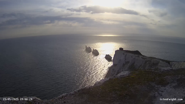 A view from a webcam showing white chalk cliffs extending into the ocean. The cliffs are topped with green grass, and a small building is visible on the edge of the highest point. The sea beneath is calm with shimmering reflections of the cloudy sky, and three large rocks are emerging from the water near the cliffs. The timestamp in the lower left corner reads "23/05/2025 19:00:24," and the website "isleofwight.com" is displayed in the lower right corner.