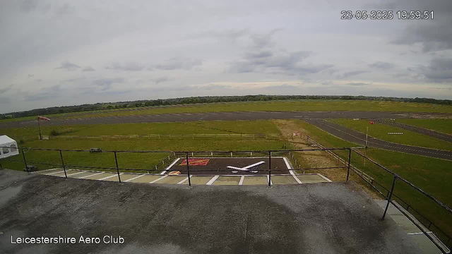 A view from a webcam showing an airfield with a small aircraft parked on a tarmac area. The runway extends into a grassy field with multiple taxiways and some distant structures under a cloudy sky. The timestamp in the top right corner indicates the date and time as 23-05-2025 at 19:59:51. The bottom left corner has text that reads "Leicestershire Aero Club."