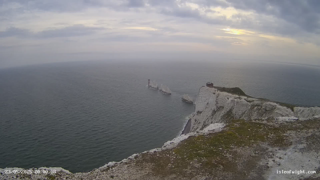 A view from a webcam showing white chalk cliffs over the sea during cloudy weather. A lighthouse or small building is on top of the cliffs, and a ship is seen navigating near the base of the cliffs in the ocean. The sky is covered with thick clouds, and the overall scene appears to be at dusk or early evening. The timestamp reads "23/05/2025 20:00:08," and the website is isleofwight.com.