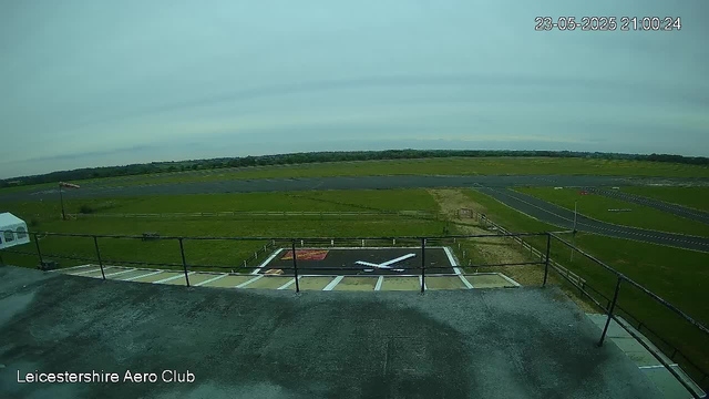 An empty helicopter landing pad with a white helicopter silhouette painted on it, surrounded by a black border. The pad is located outdoors with grassy fields extending to the horizon. In the lower-left corner, there is a logo that says "Leicestershire Aero Club." The image is from a webcam, with a timestamp in the top right corner showing "23-05-2025 21:00:24."