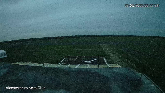 A view from a webcam showing an outdoor sports field at night. There is a helicopter landing pad with a helicopter symbol in the center. The sky appears cloudy or overcast. A small part of a road or paved area is visible in the foreground. The image has a timestamp in the top right corner reading "23-05-2025 22:00:38" and a label in the bottom left corner that says "Leicestershire Aero Club."