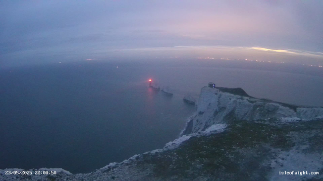 A view from a webcam showing white chalk cliffs along a coastline at dusk or dawn. The sky is partly cloudy with a pale pink and blue hue. The ocean stretches out towards the horizon, with a few small boats or ships visible. A structure is located on top of the cliff, and a red light is glowing near the edge of the water. The timestamp reads "23/05/2025 22:00:50," and the website "isleofwight.com" appears at the bottom right corner.
