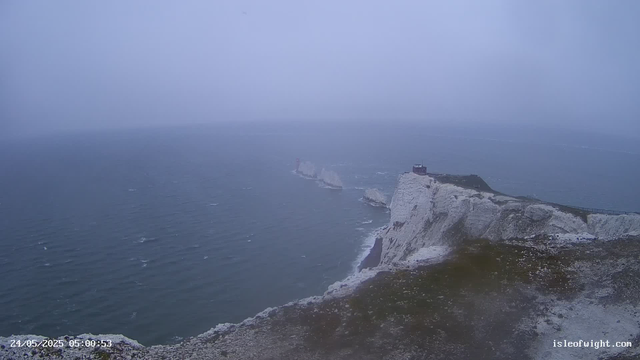 A view of white chalk cliffs rising sharply from the sea, with a lighthouse on top of the highest point. The cliffs extend into the foggy, gray ocean, which has small waves. A narrow pathway runs along the edge of the cliff. In the distance, another smaller lighthouse can be seen on a separate rocky outcrop. The sky is overcast and foggy, creating a moody atmosphere. The timestamp at the bottom left indicates the date and time as 24/05/2025 at 05:00:53, and the bottom right has the website isleofwight.com.