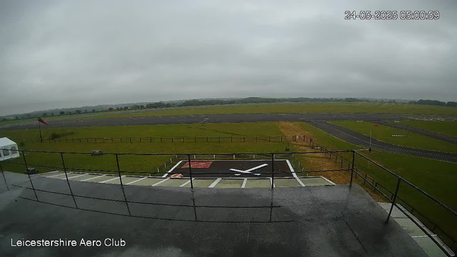 A view of an aircraft helicopter landing pad from a webcam at Leicestershire Aero Club. The scene shows a helicopter parked on the helipad, with a runway in the background under an overcast sky. The surrounding area is mostly grassy with some fencing and markings on the tarmac. The timestamp shows the date as May 24, 2025, and the time as 05:00:59.
