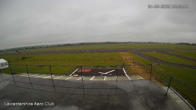 A view of an empty helicopter landing pad with a helicopter silhouette painted on it, situated in a grassy open area with a runway in the background. The sky is overcast with gray clouds. The image is timestamped "24-05-2025 05:59:58" in the top right corner. The text "Leicestershire Aero Club" is displayed in the bottom left corner. There is a small wind sock and some fencing near the landing pad. The overall scene appears to be at an airfield under cloudy weather.